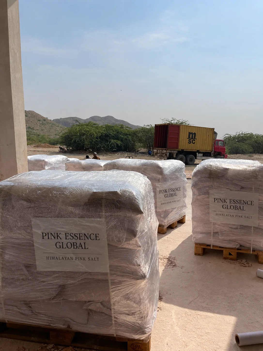 Wrapped salt bags stacked on wooden pallets in the factory yard; digitally added Pink Essence pallet labels